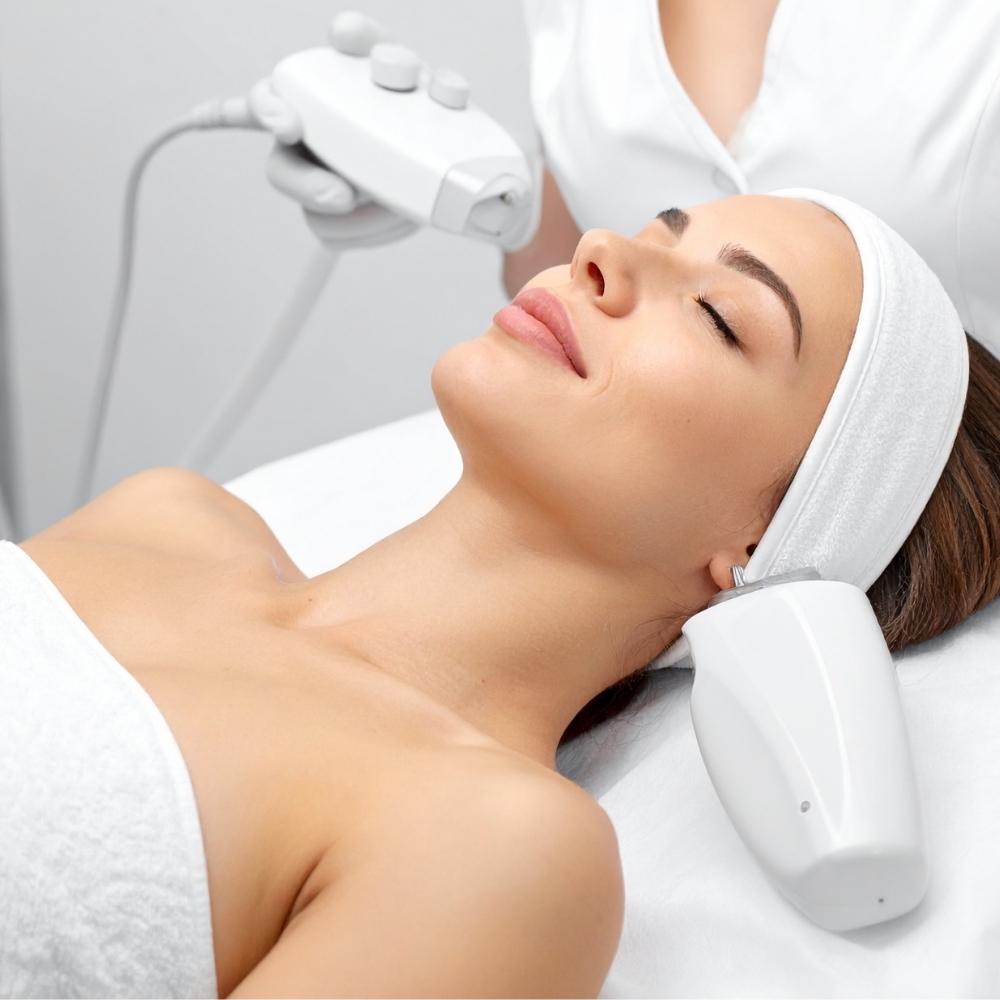 Person lying on bed wearing white towel on body and hairline, eyes closed, relaxed, another person holds a white tool near first person's face with dials and cords attached