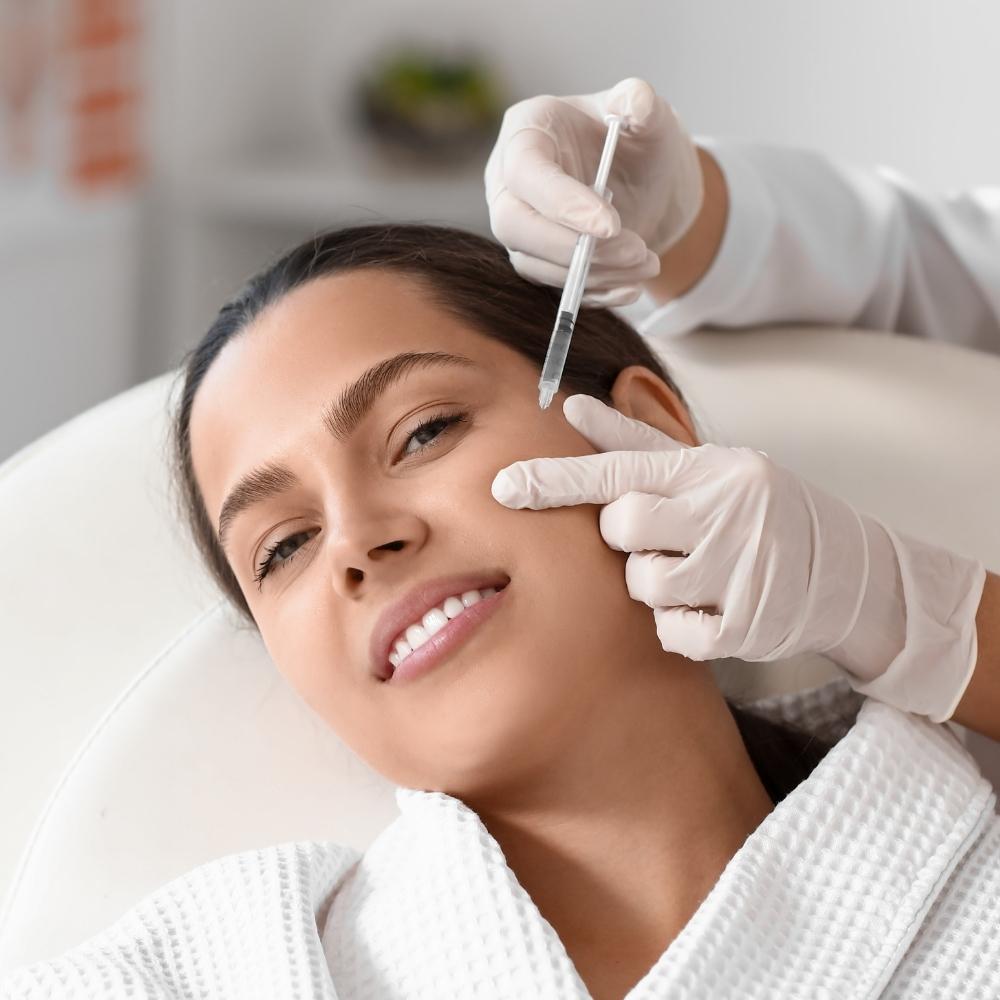 Person wearing white robe, resting head on clinical chair, looking at camera, smiling, receiving syringe injection on cheek from another person wearing white gloves