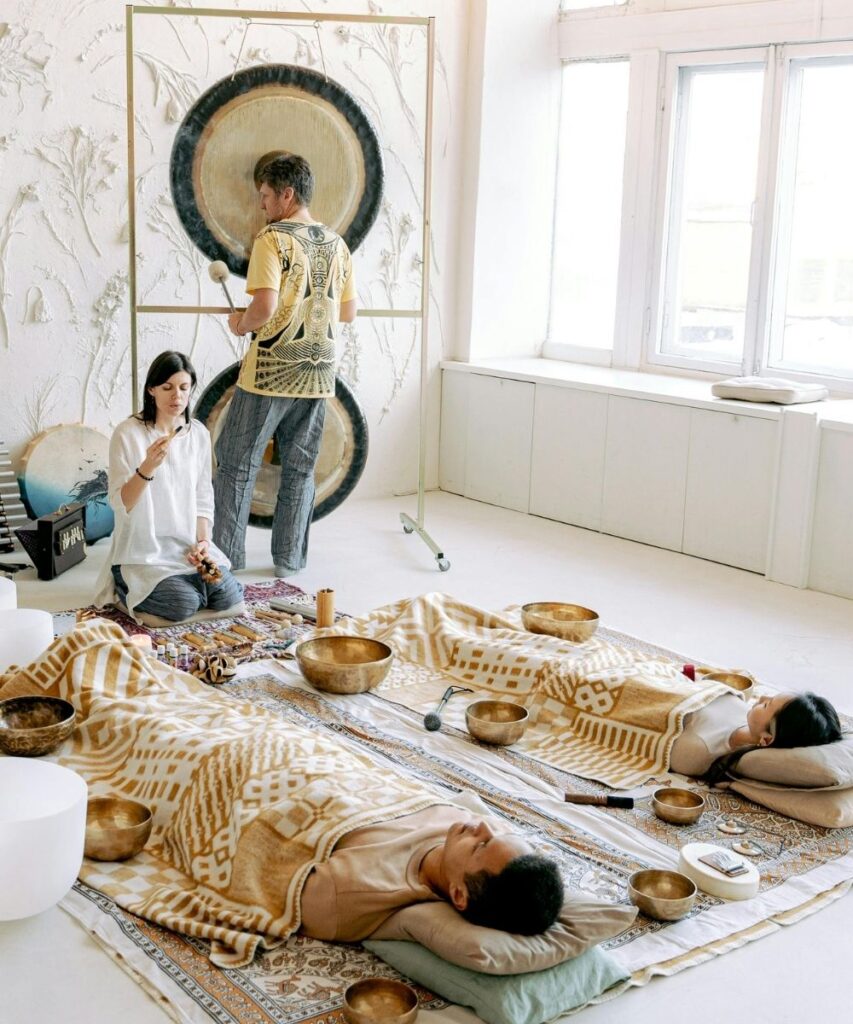 White room with four individuals, two lying on floor with rugs, pillows, blankets, and metallic bowls surrounding, one person kneeling at their feet, one person standing with mallet near gongs