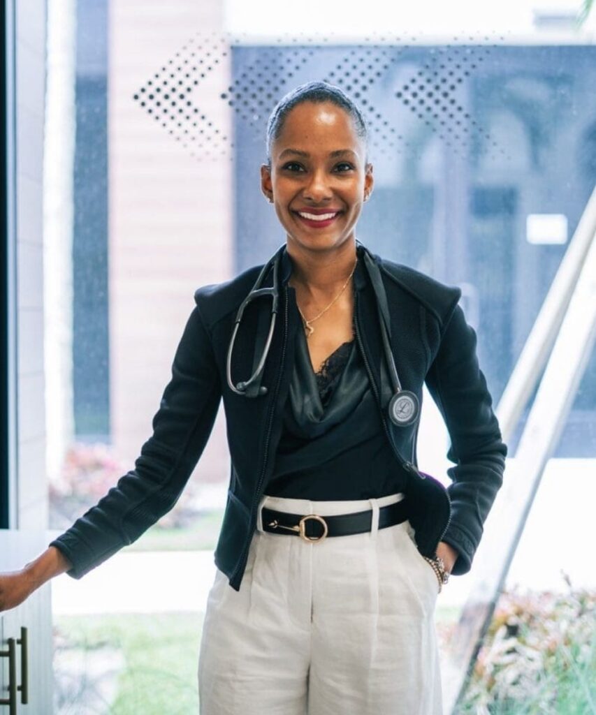 Dr. Avianne Hospedales, MD (Dr. Avi) Board-Certified Internist standing black top with black dress coat, white dress pants wearing Delray Skin Studio stethoscope